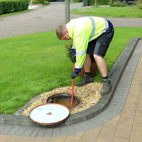A drainage expert unclogging a blocked drain on the corner of a pavement on a grass verge